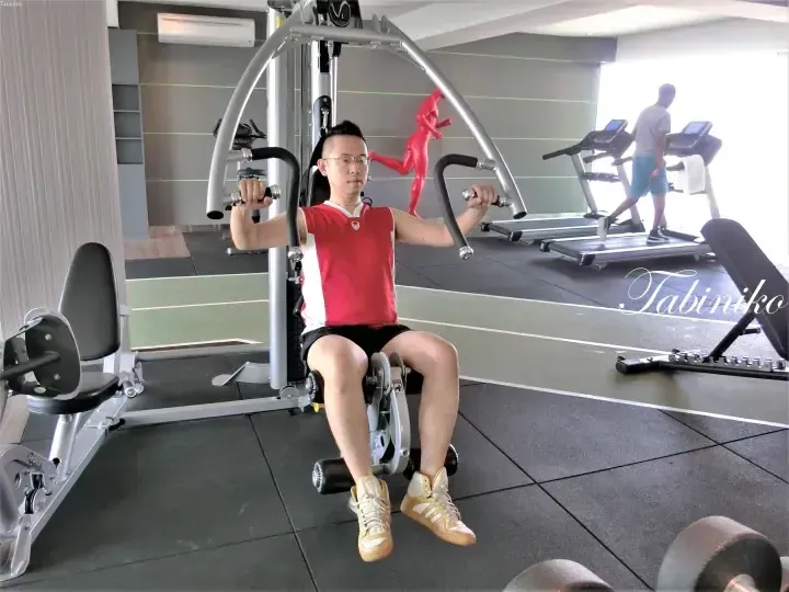 Man exercising on a chest press machine at a modern gym with treadmills and a red running figure sculpture in the background.