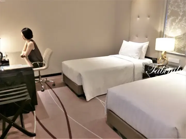 Woman sitting at a desk in a modern hotel room with two single beds and patterned carpet.