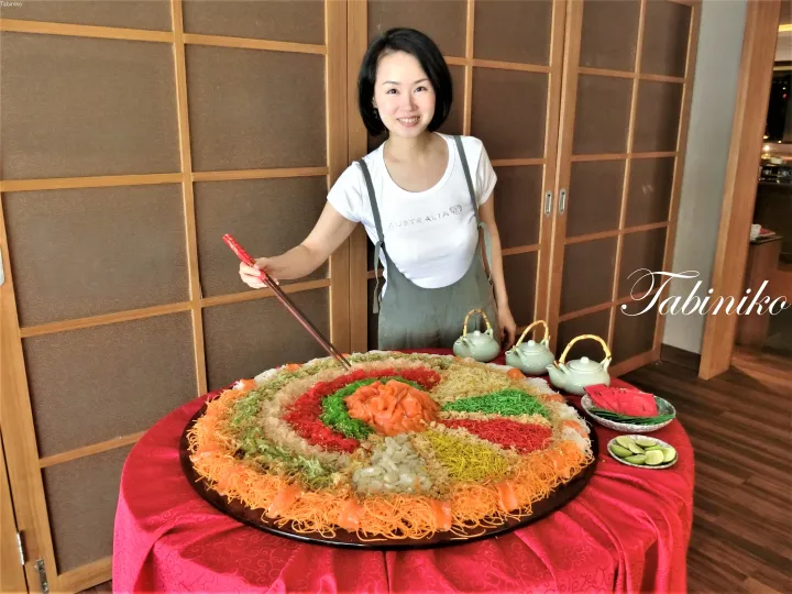 赤いテーブルクロスの上に置かれた大きな色彩豊かなユッシェン（生魚のサラダ）の前で、箸を持った女性が笑顔で写っている。