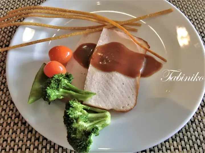 Slices of steamed fish cake with gravy, broccoli, cherry tomatoes, and crispy breadsticks on a white plate.