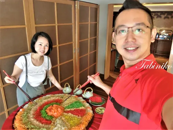 A couple smiles while holding chopsticks over a large colorful yusheng platter at a restaurant.