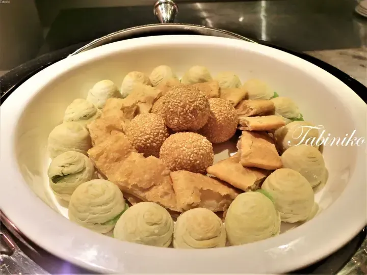 A white bowl filled with assorted pastries including sesame balls, flaky pastries, and green tea swirls at a buffet.