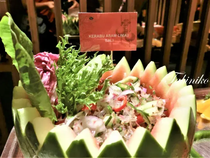 Kerabu Asam Limau Bali salad served in a carved watermelon bowl at a buffet, with a red food label.
