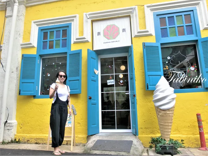 Woman holding ice cream in front of Pandan Republic&rsquo;s yellow shop with blue shutters and a giant soft serve cone statue.