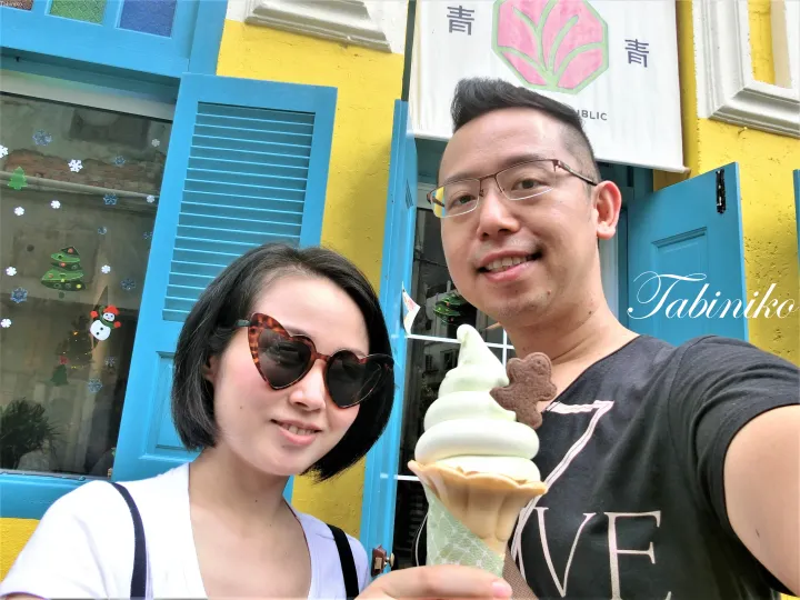 A couple posing with a green soft-serve ice cream cone in front of a yellow building with blue shutters.