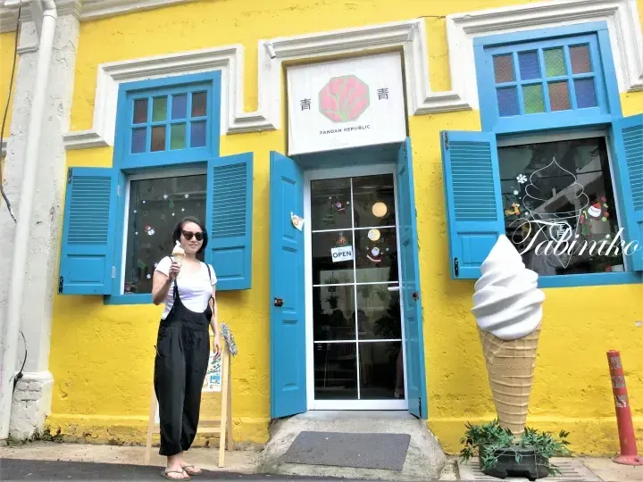 一名女子手持冰淇淋站在黄色的Pandan Republic店铺前，店铺有蓝色百叶窗和巨大的冰淇淋模型。