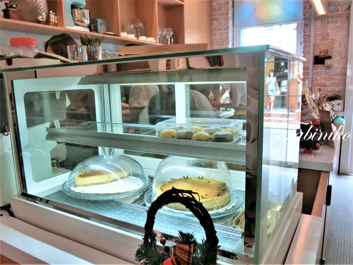 Glass display case at a cafe featuring cheesecakes under domes and pastries, with wooden shelves and brick walls in the background.