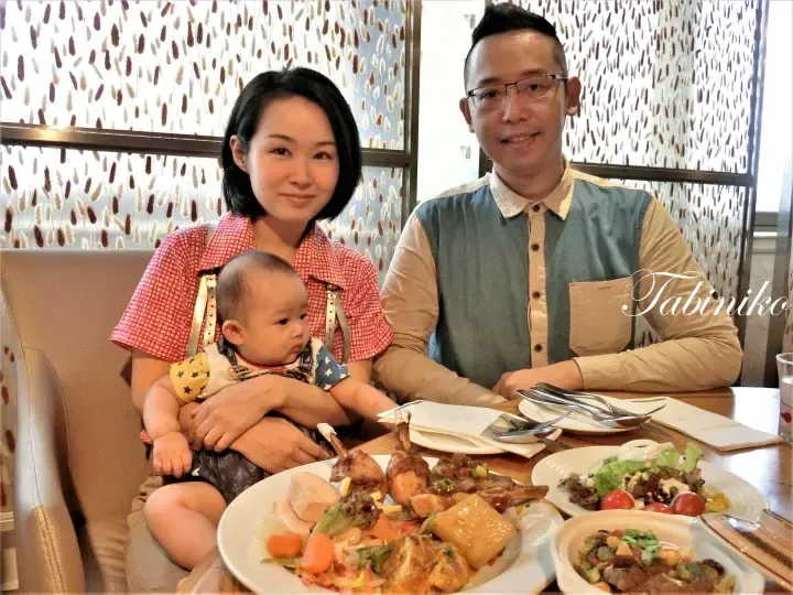 A family enjoys a meal at a restaurant with patterned walls, featuring plates of roasted lamb chops and fresh salad.