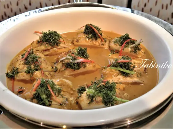 Grilled chicken served in a creamy yellow curry sauce, garnished with crispy herbs and julienned vegetables in a white bowl.