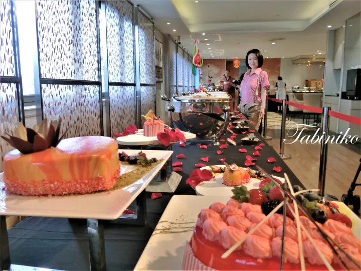 光沢のあるオレンジ色のケーキやピンクのパステリー、バラの花びらが飾られたビュッフェのデザートテーブル。