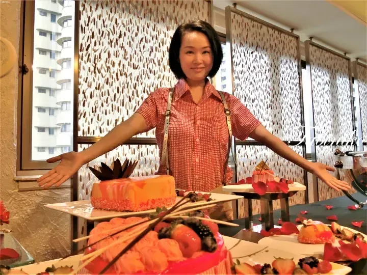 Smiling woman in red shirt presenting a dessert buffet display with cakes and fruit skewers at a hotel.