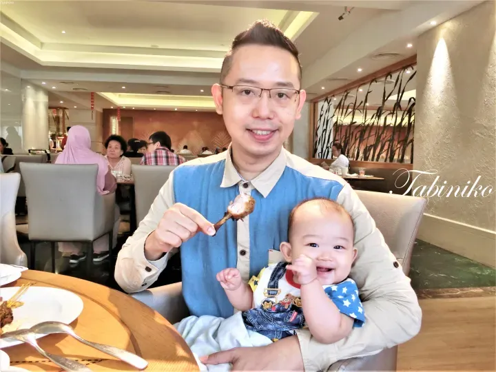一名男子在餐廳裡抱著嬰兒，手裡拿著一塊沙嗲微笑著。