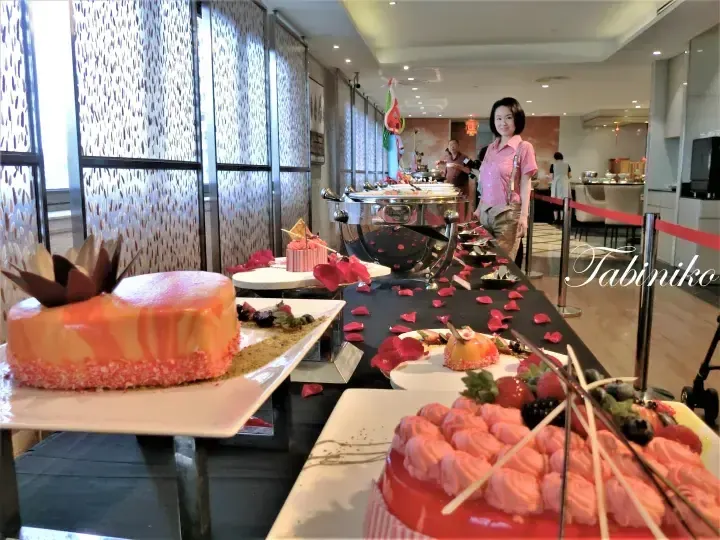 Buffet dessert table featuring a glossy orange cake and pink pastries with rose petals, with a woman standing nearby.
