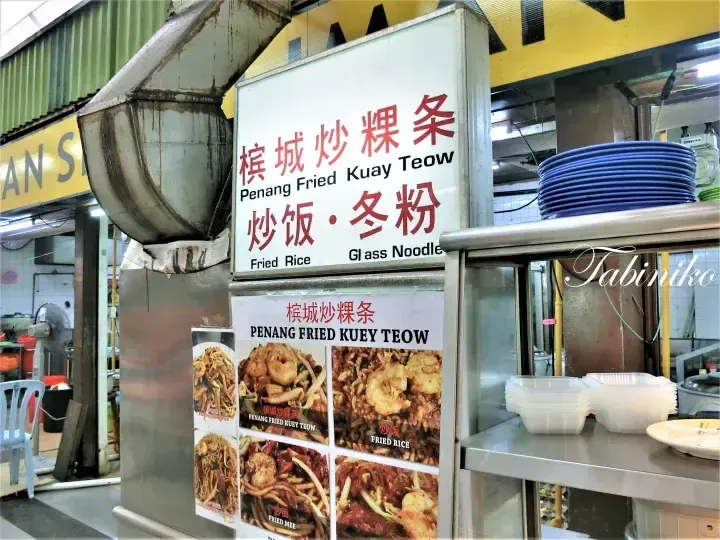 Menu board at a Malaysian hawker stall advertising Penang Fried Kuay Teow, fried rice, and glass noodles with photos of the dishes.