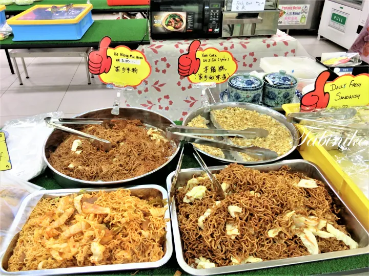 黄色い看板が並ぶマレーシアのハワーカーで、カヒョンミーフーンや芋頭糕など様々な料理が金属製のトレイに並んでいる。