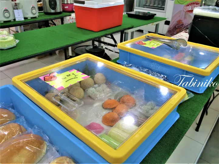 セルフサービスフードスタールの緑色のテーブルに、クイやパンなどの伝統的なマレーシアのスナックがカラフルなプラスチック容器に入っている。