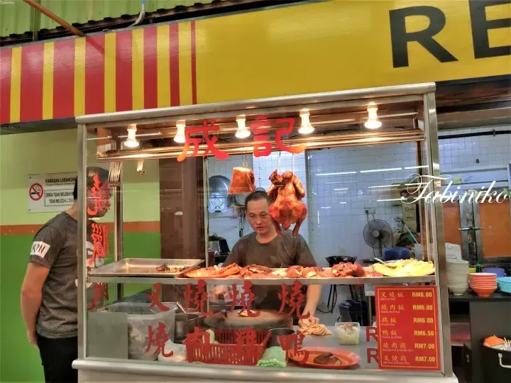 A vendor at Cheng Kee stall prepares roasted meats including BBQ pork and chicken, with a menu displaying prices in Malaysian Ringgit.