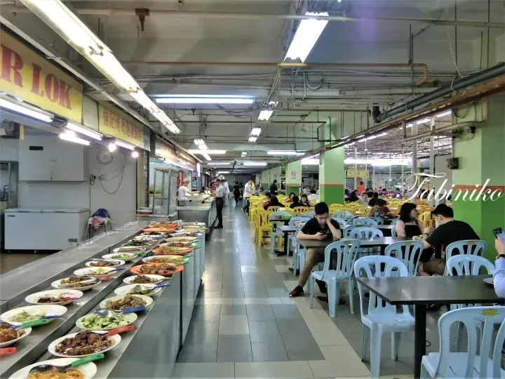 Long buffet counter displaying various dishes at a Malaysian food court with diners seated at tables.