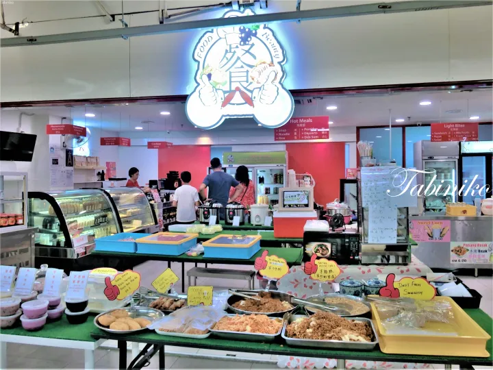 照明サイン、陳列ケース、ホットミールや軽食を注文する客が映るフードスタールの店内。