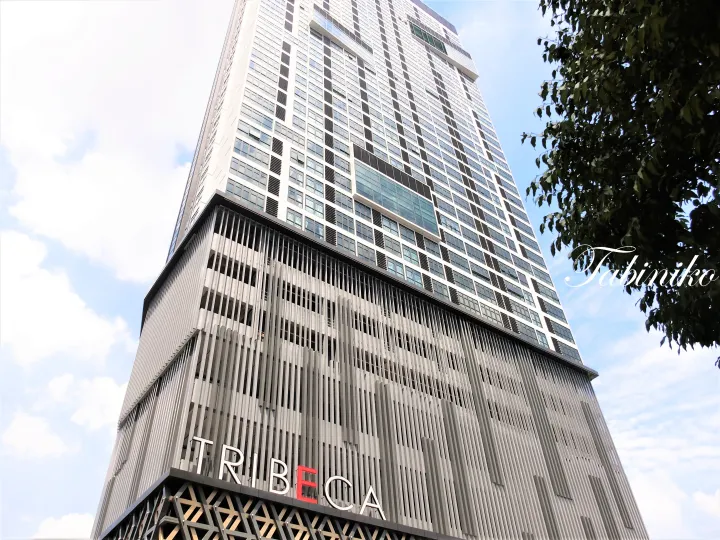 青空を背景に、外壁に垂直の格子状デザインと「Tribeca」の看板が目立つモダンな高層ビル
