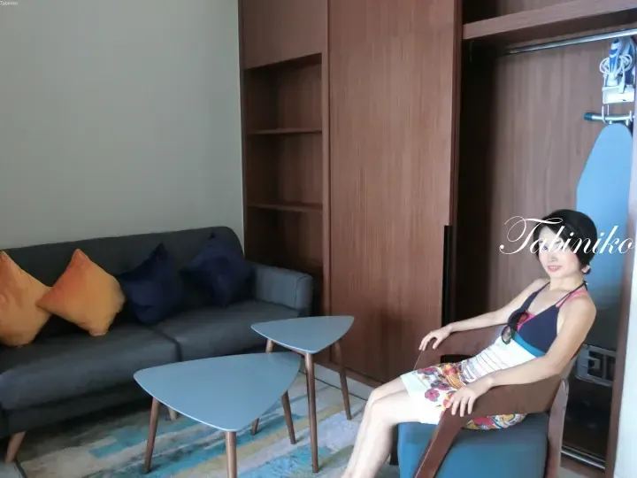 Woman relaxing in a modern hotel suite living area with a grey sofa, wooden wardrobe, and nesting coffee tables.