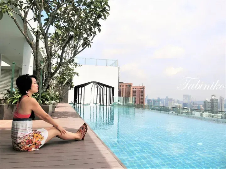 Woman relaxing by an infinity pool with a view of the Kuala Lumpur skyline, including the Menara Maybank building.