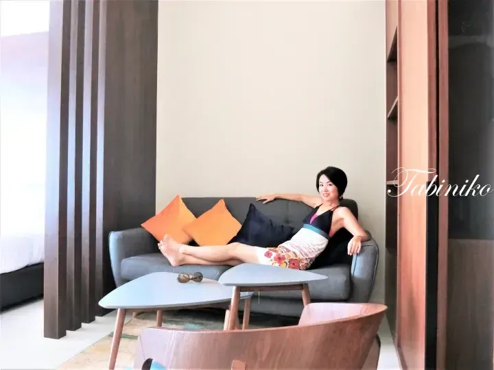 Woman relaxing on a grey sofa in a modern hotel suite living area with orange cushions and a wooden coffee table.
