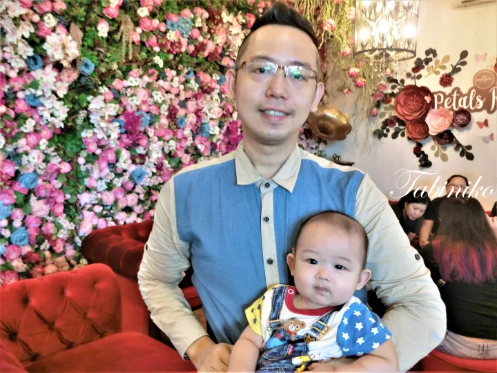 マレーシアのPetals Roomカフェで花の壁の前で赤ん坊を抱く男性。