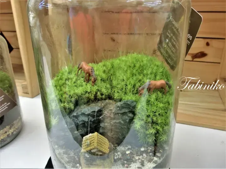 Terrarium with miniature horses on mossy terrain and a small house, displayed in a glass jar.