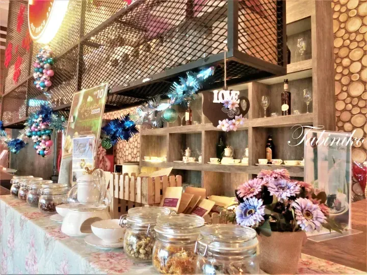 Cafe counter with glass jars of tea, floral tablecloth, and wooden shelves displaying ‘LOVE’ sign and glassware.
