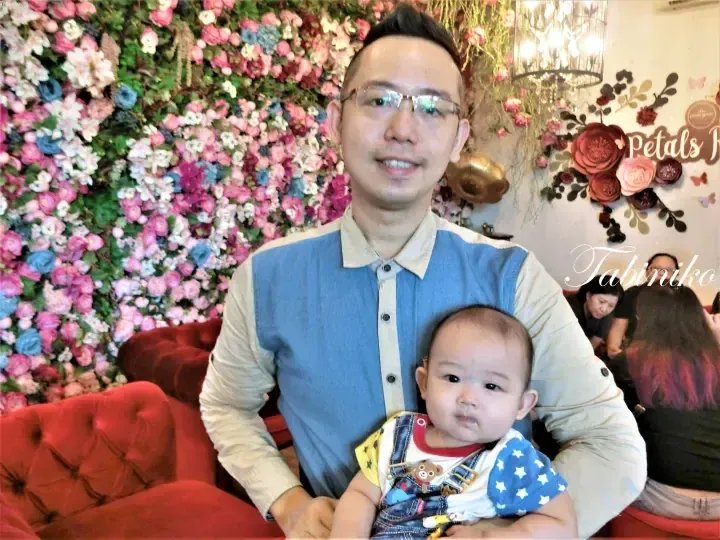 Man holding a baby in front of a floral wall at Petals Room cafe in Malaysia.