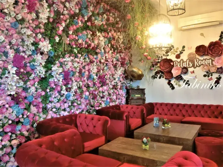 A cozy lounge area with plush red velvet sofas and wooden tables set against a vibrant floral wall at Petals Room.