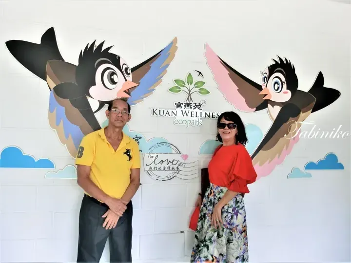 A couple posing in front of a wall mural featuring cartoon swallows and the Kuan Wellness ecopark logo.