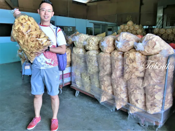 大量のポテチやスナック菓子が積まれた倉庫で、エビせんべいの大きな袋を持つ男性。