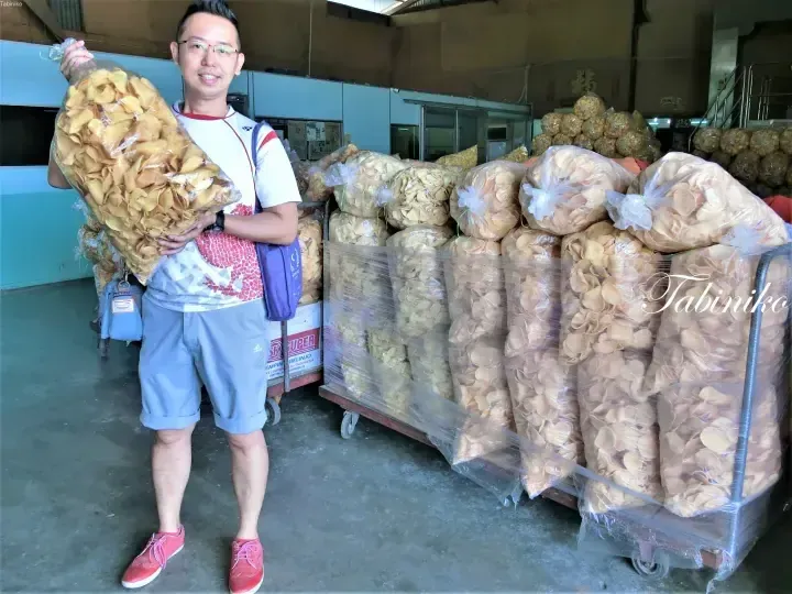 Man holding a large bag of prawn crackers in a warehouse filled with bulk snack packages.