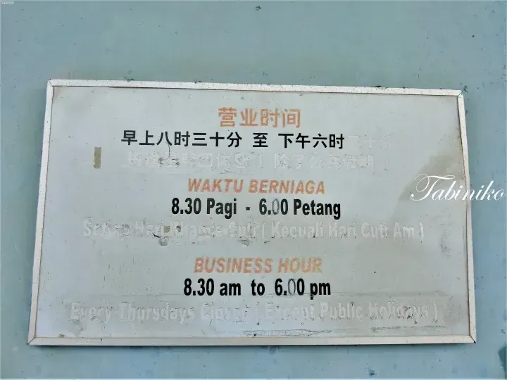 Weathered sign displaying business hours in Chinese, Malay, and English, indicating 8:30 am to 6:00 pm with Thursday closures.