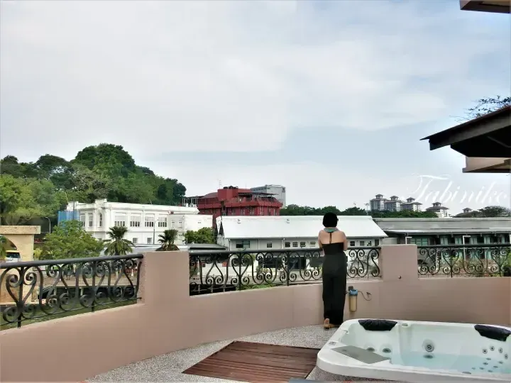 Woman in black outfit stands on a balcony with a hot tub overlooking the Malacca River and colonial buildings.