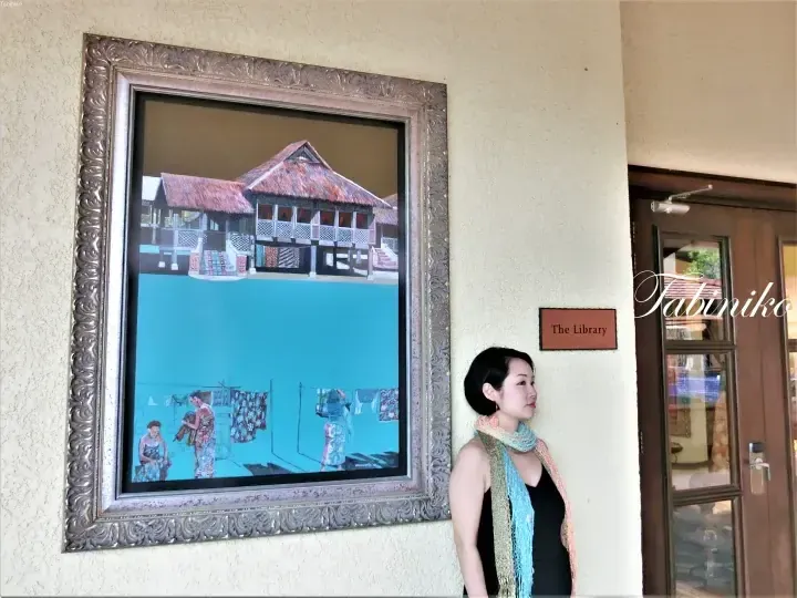 Woman stands beside a framed painting of a traditional stilt house and laundry scene outside The Library entrance.