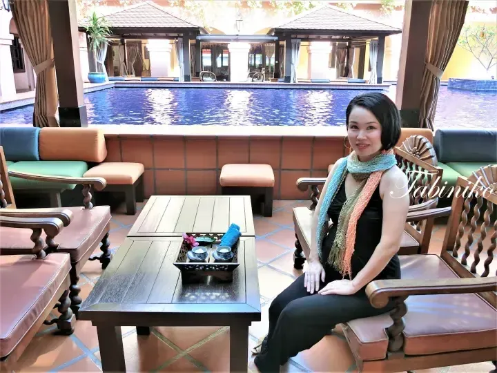 Woman sitting on wooden chair at poolside lounge with tea tray, overlooking resort pool and pavilions.