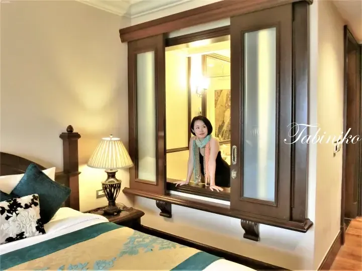 Woman leaning through a wooden sliding window connecting a bedroom to a bathroom in a luxury hotel suite.