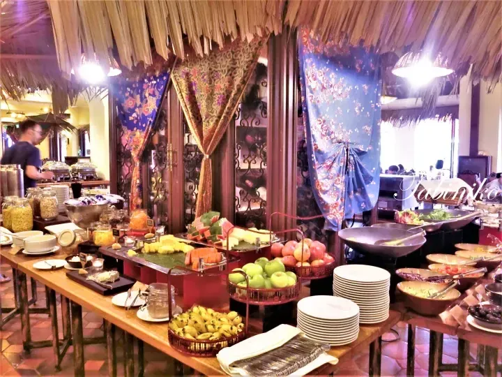 Buffet spread featuring fresh fruits, cereals, and hot dishes under thatched roofing at a Malaysian resort dining area.