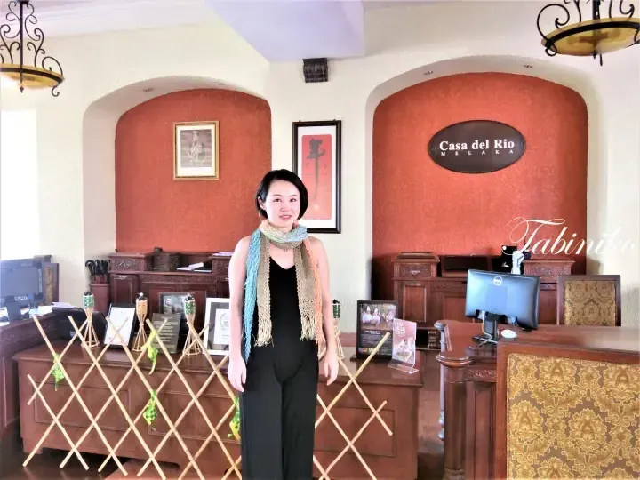 Woman standing at the reception desk of Casa del Rio Melaka hotel lobby with wooden furniture and red walls.