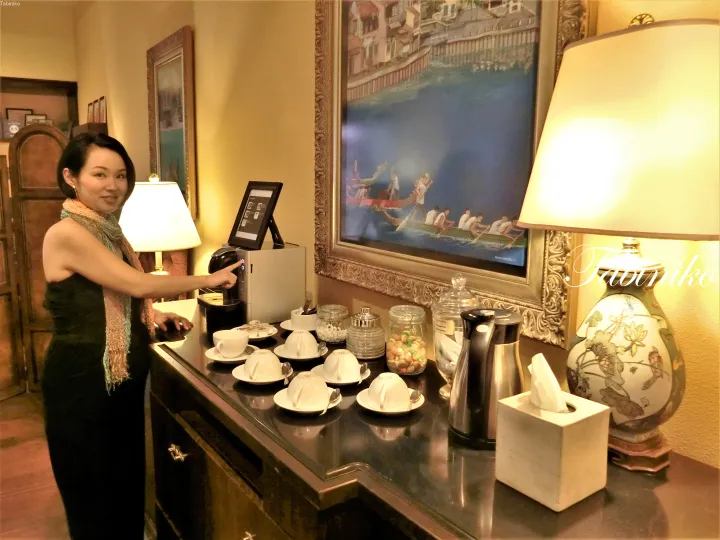 コーヒーメーカーを使用する女性、コーヒーセットと龍舟の絵画が飾られたホテルルーム。