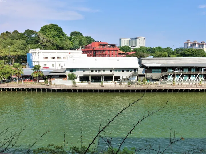ジョホールバルルの木製桟橋沿いにある白と赤の建物と、背景の現代的なホテルの川沿いの景色。
