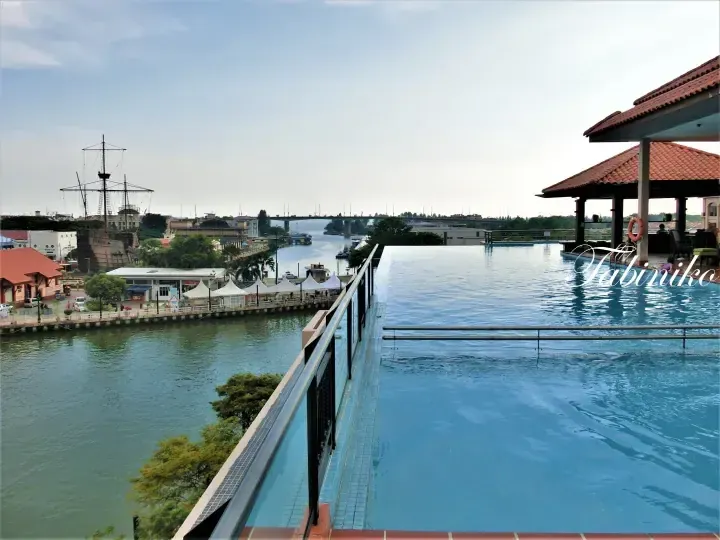 Infinity pool overlooking Melaka River with a pirate ship replica and Melaka Bridge in the background.
