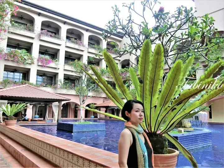 Woman standing by a pool with large bird&rsquo;s nest ferns at a resort featuring balconies with bougainvillea.