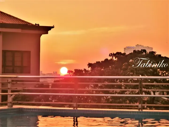 Sunset view over a swimming pool and balcony railing with a building and trees silhouetted against the orange sky.