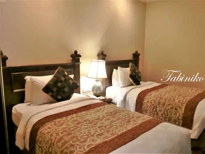 Two twin beds with dark wood headboards and patterned runners in a warmly lit hotel room.