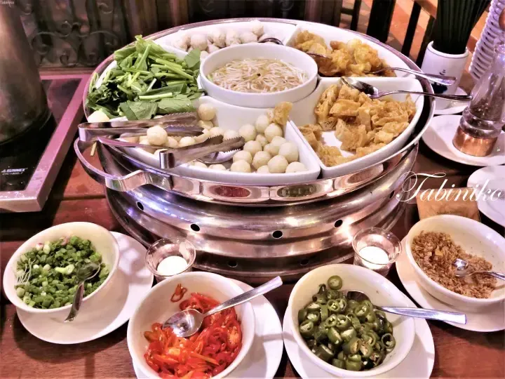Buffet spread featuring a tiered hot pot station with meatballs, greens, noodles, and fried tofu alongside bowls of fresh chilies and herbs.