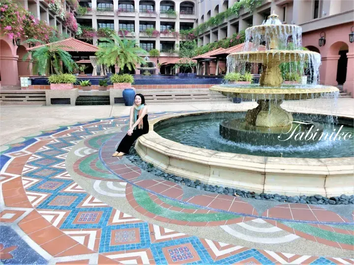 Woman sitting beside a tiered fountain in the courtyard of The Datai Langkawi, surrounded by intricate mosaic tiles and lush tropical greenery.